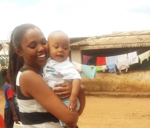 Chimwemwe smiles holding a baby in front of a building and clothesline.